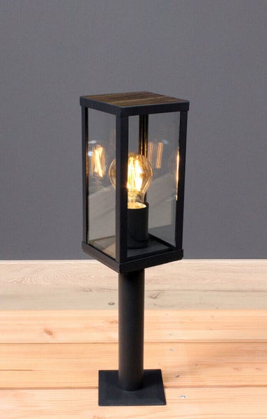 Moderne schwarze Außen-Sockelleuchte mit rechteckiger Glaslaterne, Abdeckung in Holzoptik und sichtbarem Filament-Leuchtmittel auf einem Metallpfosten.