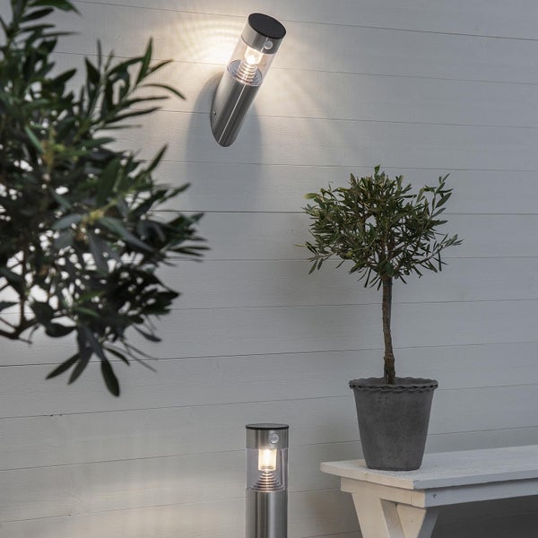 Szene mit einer Wandleuchte und einer Wegeleuchte aus Metall auf einer Holzterrasse mit Pflanzen