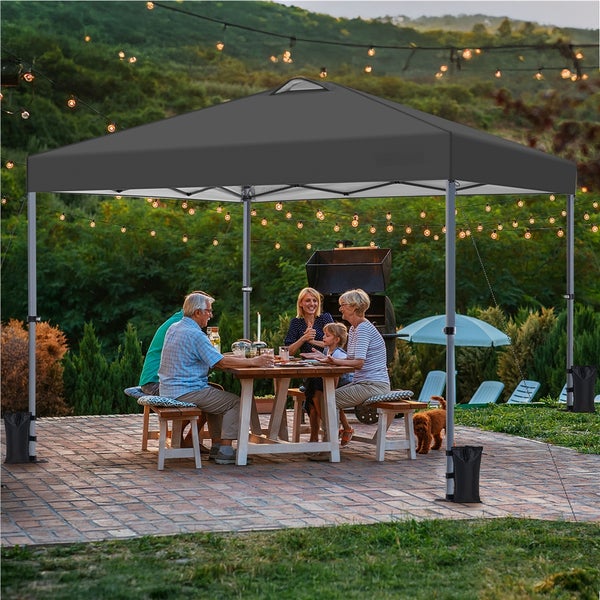 Szene im Freien mit einem grauen Pavillon, unter dem Menschen an einem Holztisch sitzen, mit Dekorationen und einer entspannten Atmosphäre