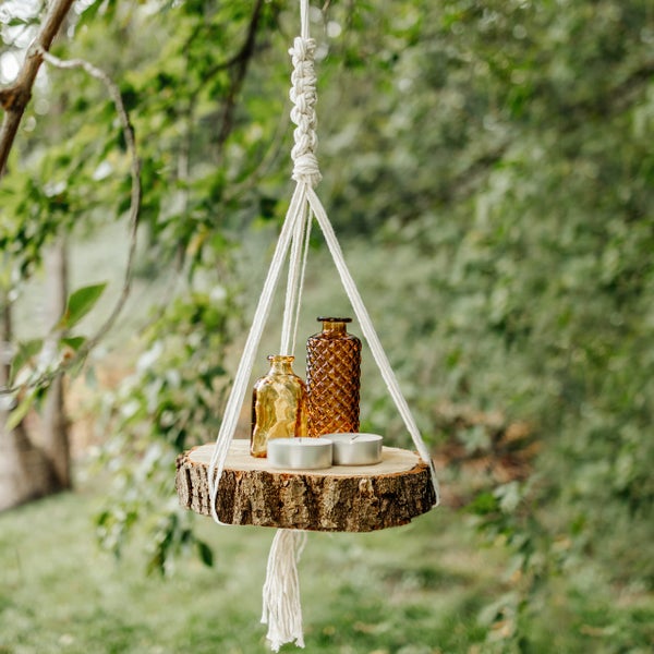 Dekoratives Hängeregal aus einer Holzscheibe mit Makramee-Seil, bestückt mit Glasflaschen und Teelichtern in einem hellen Garten.