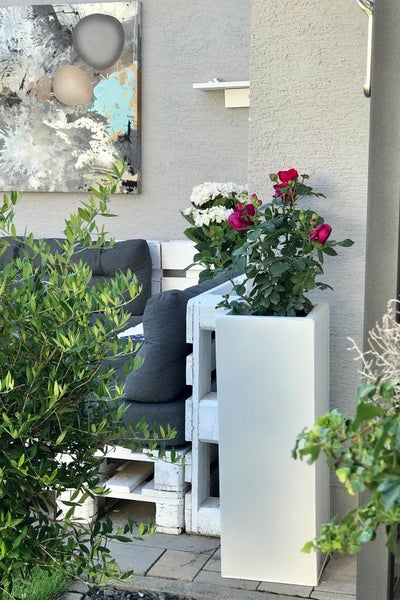 Hoher weißer Pflanzkübel mit Rosen und Hortensien neben einem weißen Palettensofa mit grauen Polstern auf einer Terrasse.
