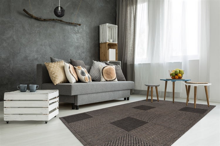 Wohnzimmer mit grauem Sofa, strukturierten Kissen, Holzkisten als Regale und Beistelltische auf einem modernen, dunkel gemusterten Webteppich.