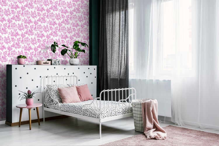 Helles Schlafzimmer mit weißem Metallbett, gemusterter Bettwäsche und rosa Akzenten vor einer Wand mit pinkfarbener Tapete.