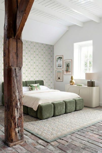 Helles Schlafzimmer mit grünem Polsterbett, Tapete mit Sonnenmotiven, rustikalem Holzbalken und gemustertem Teppich auf Backsteinboden.