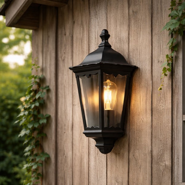 Schwarze Außenwandleuchte im Laternenstil mit Glaseinsätzen und leuchtendem Leuchtmittel an einer Holzwand mit Rankpflanzen.