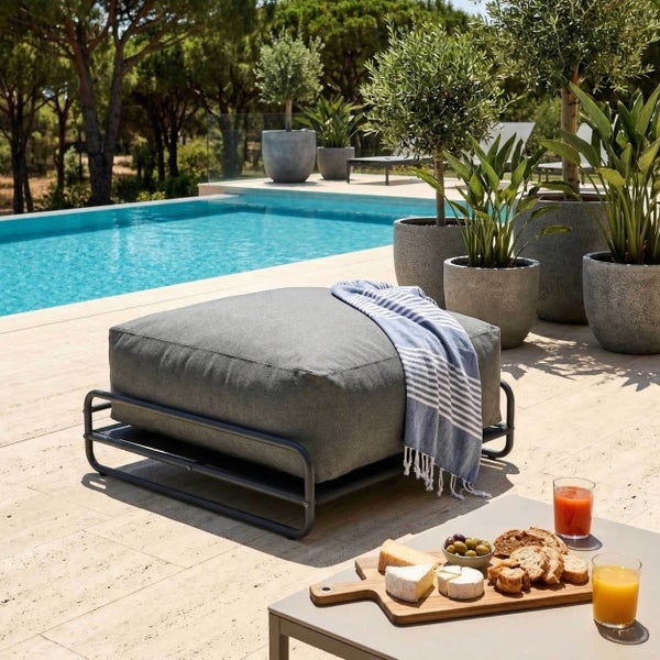 Moderner Gartenhocker mit grauem Polster und schwarzem Metallrahmen auf einer sonnigen Terrasse am Pool, umgeben von Pflanzen.