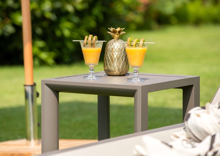 Ein quadratischer Beistelltisch aus Metall steht im Garten, darauf zwei Gläser mit Saft und ein Deko Objekt in Ananasform.