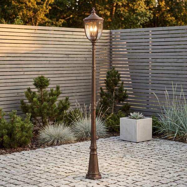 Klassische Wegeleuchte aus Metall auf gepflasterter Terrasse vor einem Holzzaun mit Pflanzen im Garten.