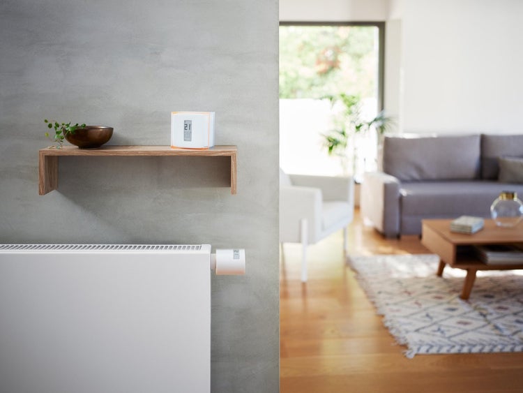 Smarte Heizungssteuerung mit Raumthermostat auf einem Holzregal und elektronischem Heizkörperthermostat an einem weißen Heizkörper im Wohnzimmer.