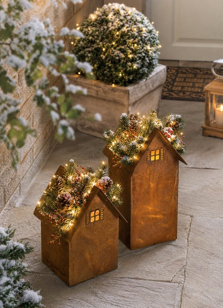 Zwei dekorative Metallhäuser in Edelrostoptik mit weihnachtlicher LED Beleuchtung, Tannenzweigen und Zapfen für die Außendekoration.