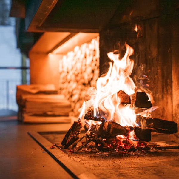 Brennendes Kaminfeuer mit Holzscheiten in einem offenen Kamin mit Kaminholzlager im Hintergrund