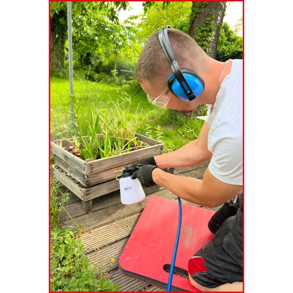 Mann sprüht Pflanzen in einem Holzkasten mit einer Gartenspritze, während er Knieschoner, Gehörschutz und Schutzbrille trägt.