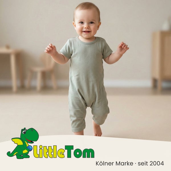 Ein lächelndes Baby steht in einem hellgrünen Einteiler in einem hellen Zimmer mit Holzmöbeln. LittleTom Logo.
