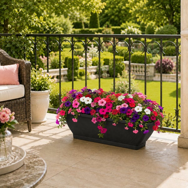 Ein länglicher schwarzer Blumenkasten mit bunten Petunien auf einem Balkon mit Blick in einen Garten.