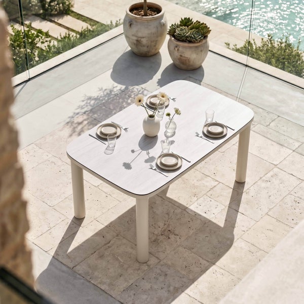 Moderner Gartentisch mit abgerundeten Ecken, helles Design, vier dicke Beine, eingedeckt auf einer Steinterrasse neben einem Pool.