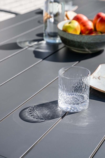 Strukturiertes Trinkglas auf einem Gartentisch mit Lattenstruktur im hellen Sonnenlicht mit Lichtreflexionen.