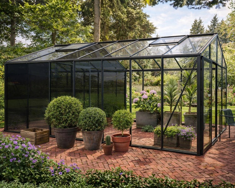 Großes Gewächshaus mit schwarzem Metallrahmen auf einer Ziegelterrasse, bepflanzt mit Topfpflanzen in einem hellen, organisierten Garten.