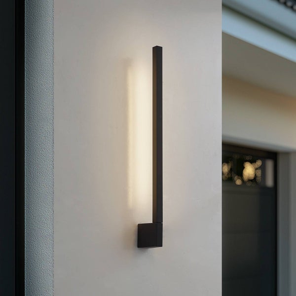 Moderne schwarze Außenwandleuchte in vertikaler Stabform mit warmweißem indirektem Licht an einer Wand.