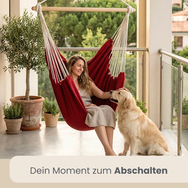 Frau entspannt in einem roten Hängesessel mit Holzstab auf einem hellen Balkon mit ihrem Hund und verschiedenen Topfpflanzen.