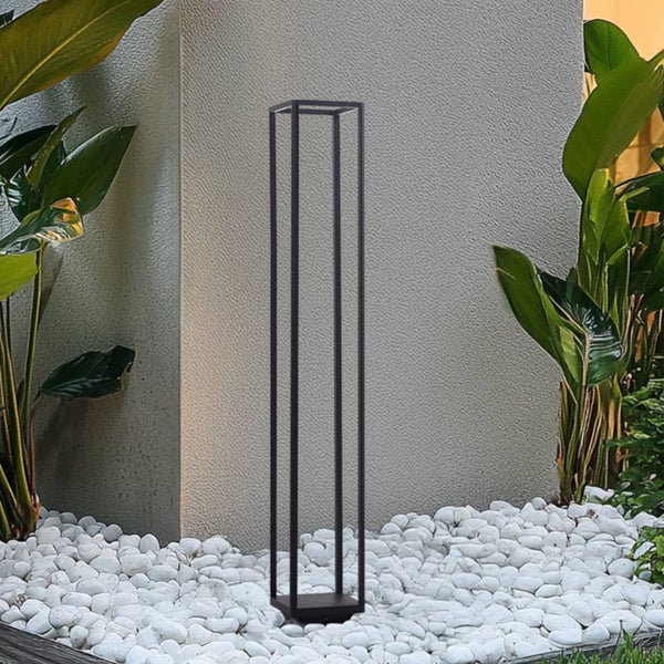 Schwarze minimalistische Außenstehleuchte in rechteckiger Rahmenform auf weißen Kieselsteinen neben grünen Pflanzen vor einer grauen Wand.