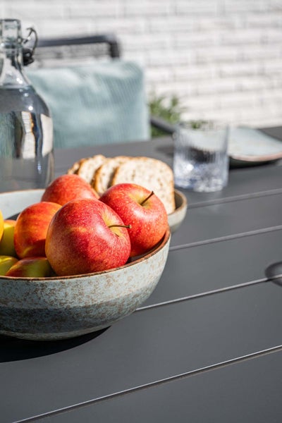 Dunkelgrauer Gartentisch mit Lattenstruktur, dekoriert mit einer Schale roter Äpfel, Brot und einer Wasserflasche in einer hellen Außenumgebung.