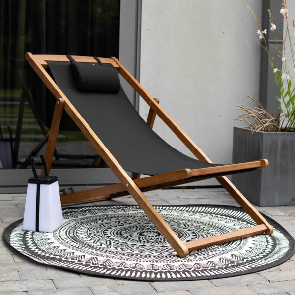 Liegestuhl aus Holz mit schwarzer Stoffbespannung und Kopfkissen auf einem gemusterten Outdoor-Teppich neben einer tragbaren Leuchte.