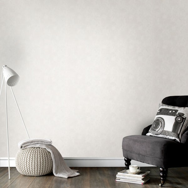 Wohnzimmer mit unifarbener Vliestapete, Sessel, Stehlampe, Strickhocker mit Decke und Stapel Zeitschriften vor einer schlichten Wand.