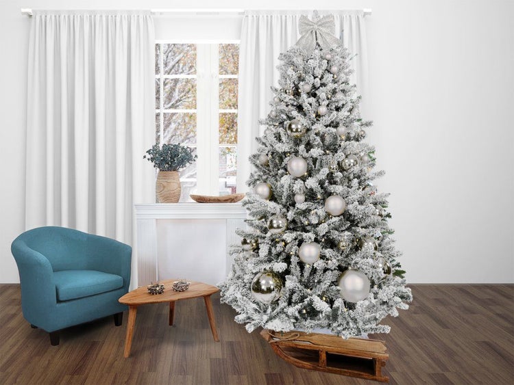 Ein künstlicher, beschneiter Weihnachtsbaum mit Kugeln und LED Beleuchtung steht in einem Wohnraum vor einem Fenster mit einem Holzschlitten.