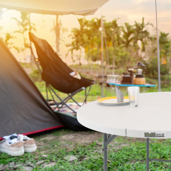 Runder Klapptisch von Trutzholm auf einer Wiese vor einem Zelt mit Campingstuhl und Ausrüstung im Hintergrund bei Sonnenuntergang.