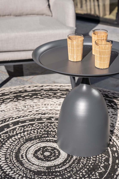 Moderner dunkelgrauer Beistelltisch aus Metall mit runder Tischplatte und glockenförmigem Fuß auf einem gemusterten Outdoor-Teppich.