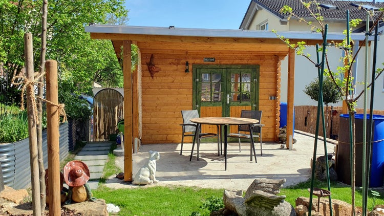 Gartenhaus aus Holz mit Flachdach, Doppeltür und gepflasterter Terrasse mit Tisch und Stühlen in einem hellen Garten.