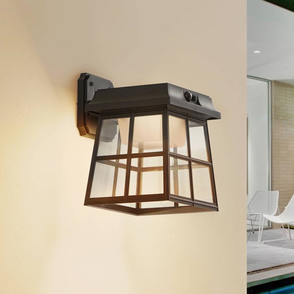 Beleuchtete schwarze Außenwandleuchte im Laternen-Design mit klaren Glasscheiben und Gitterstruktur, montiert an einer hellen Wand.