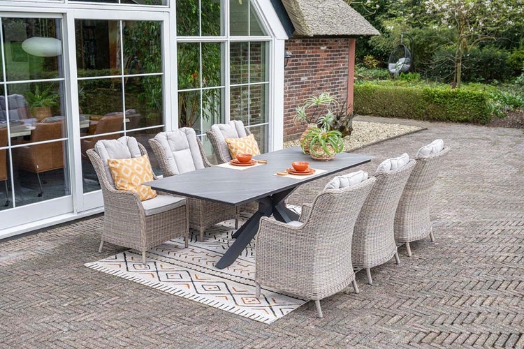 Garten-Essgruppe mit großem grauem Tisch, sechs Rattansesseln mit Polstern und einem gemusterten Außenteppich auf einer Terrasse.