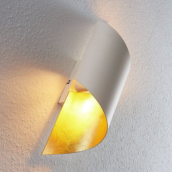 Wandleuchte in geschwungenem Design, weiße Außenseite, goldfarbene Innenseite, warmes Licht auf strukturierter Wand.