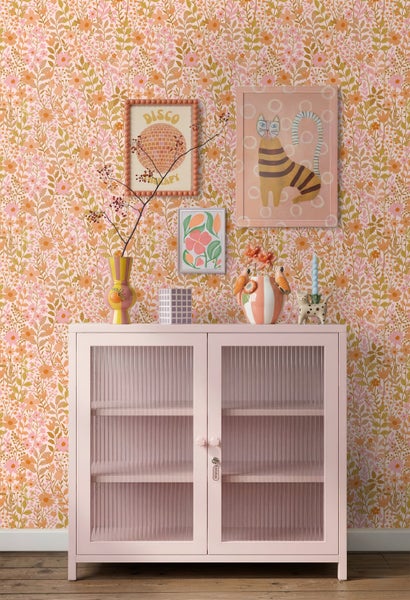 Vliestapete mit floralem Muster in Orange und Rosa hinter einem rosa Schrank mit Riffelglastüren und bunter Dekoration.