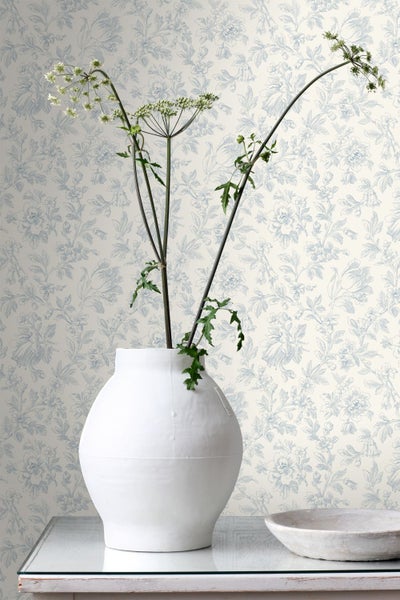 Vliestapete mit floralem Muster in Blau auf hellem Hintergrund, kombiniert mit einer weißen Vase und getrockneten Pflanzen.