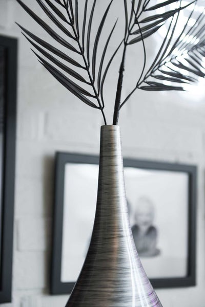 Moderne Vase aus gebürstetem Metall mit künstlichen Palmzweigen vor einer Wand mit Bilderrahmen.