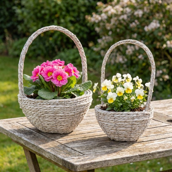 Zwei geflochtene Pflanzkörbe mit Henkel stehen auf einem Holztisch, bepflanzt mit rosa Primeln und weiß gelben Stiefmütterchen im Garten.