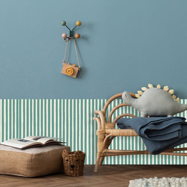 Kinderzimmer mit Rattan-Stuhl, Kissen, Decke und Spielzeug vor einer Wand mit Streifenmuster.