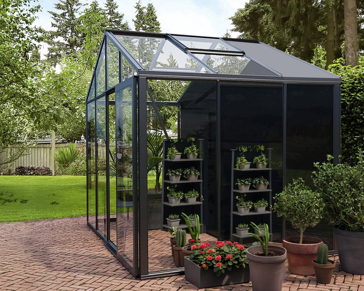 Modernes Gewächshaus aus Glas und Metall auf einer Terrasse, ausgestattet mit Pflanzenregalen und Dachfenster, in einem organisierten Garten.