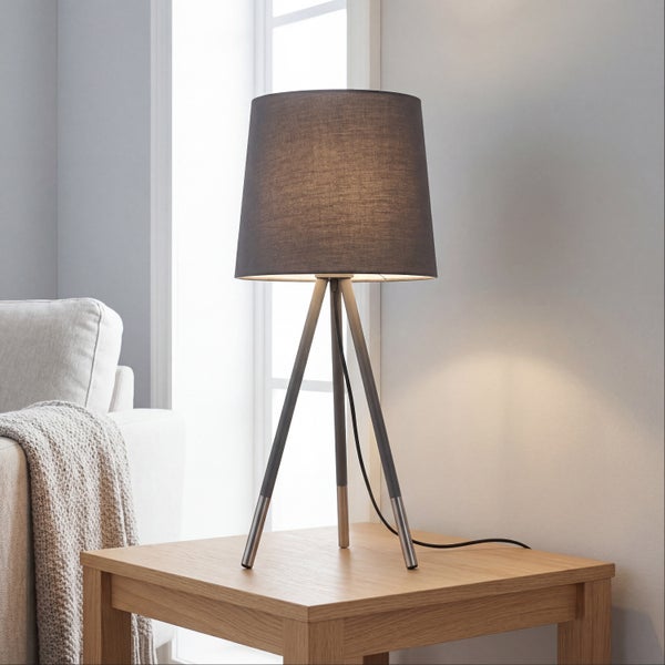 Moderne Tischleuchte mit dreibeinigem Metallgestell und konischem Lampenschirm auf einem Holztisch in einem Wohnraum.