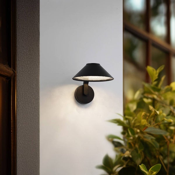 Moderne schwarze Außenwandleuchte mit kegelförmigem Schirm, montiert an einer hellen Hauswand neben Pflanzen.