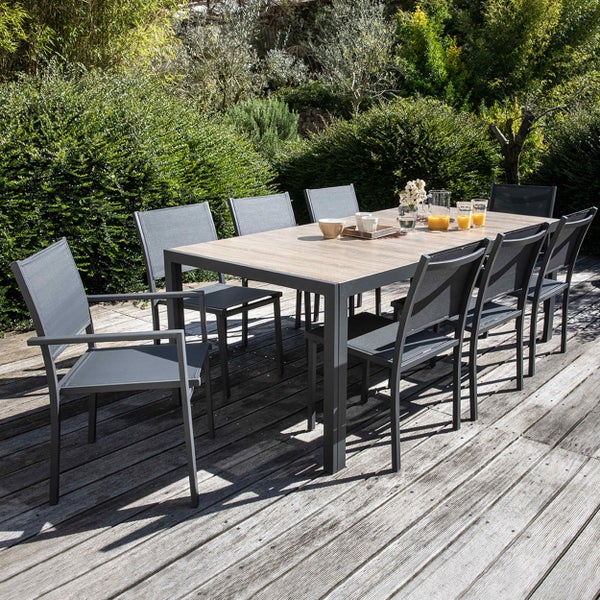 Garten-Essgruppe auf Holzterrasse mit rechteckigem Holztisch und acht grauen Metallstühlen mit Textilbespannung vor grüner Hecke.