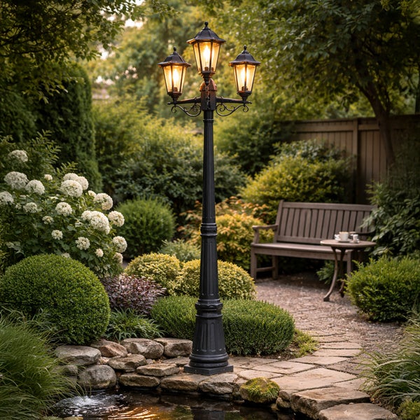 Dreiflammige Gartenmastleuchte im klassischen Design stehend an einem Teich in einem gepflegten Garten mit Sitzbank und Blumenbeeten.