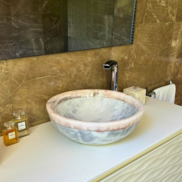 Modernes Badezimmer mit einem runden Aufsatzwaschbecken aus Naturstein auf einem weißen Waschtisch vor einer Wand aus braunem Marmor.