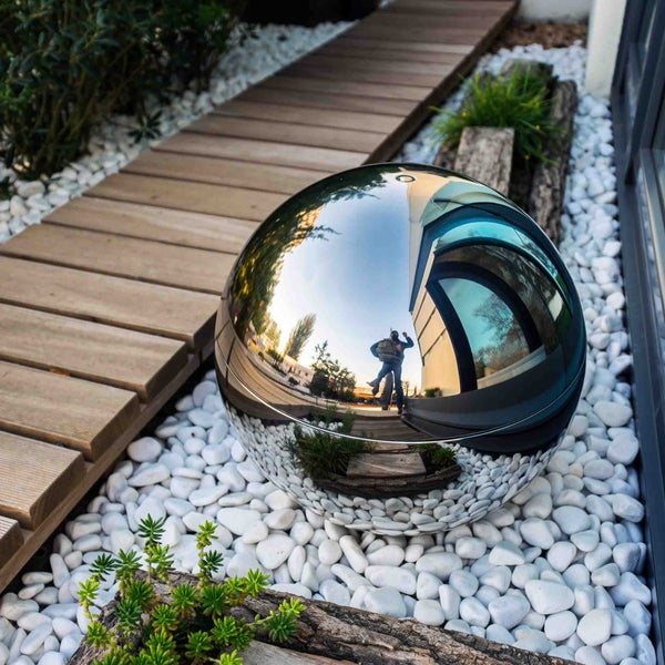 Reflektierende Gartenkugel aus Edelstahl auf weißen Kieselsteinen neben einem Holzweg im modernen Garten.
