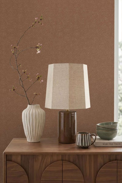 Wohnraum mit brauner Tapete mit Fischgrätenmuster, Tischlampe auf einem Holzschrank, eine Vase mit Zweigen und Keramikschalen.