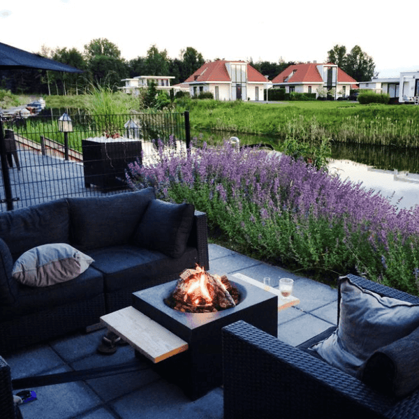 Moderne Terrasse mit quadratischer Feuerschale und dunklem Lounge-Set an einem Kanal in der Dämmerung.
