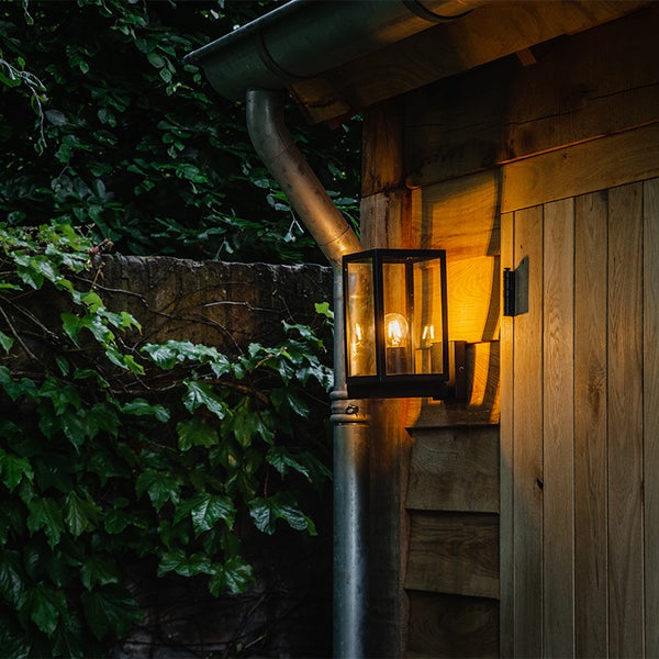 Moderne schwarze Außenwandleuchte mit Glaseinsätzen und leuchtendem Leuchtmittel an einer Gartenhütte aus Holz neben einem Fallrohr