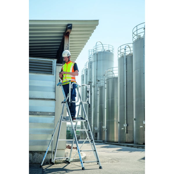 Eine Person mit Schutzhelm und Warnweste steht auf einer Zarges Treppenleiter vor einem Gebäude mit Tanks.